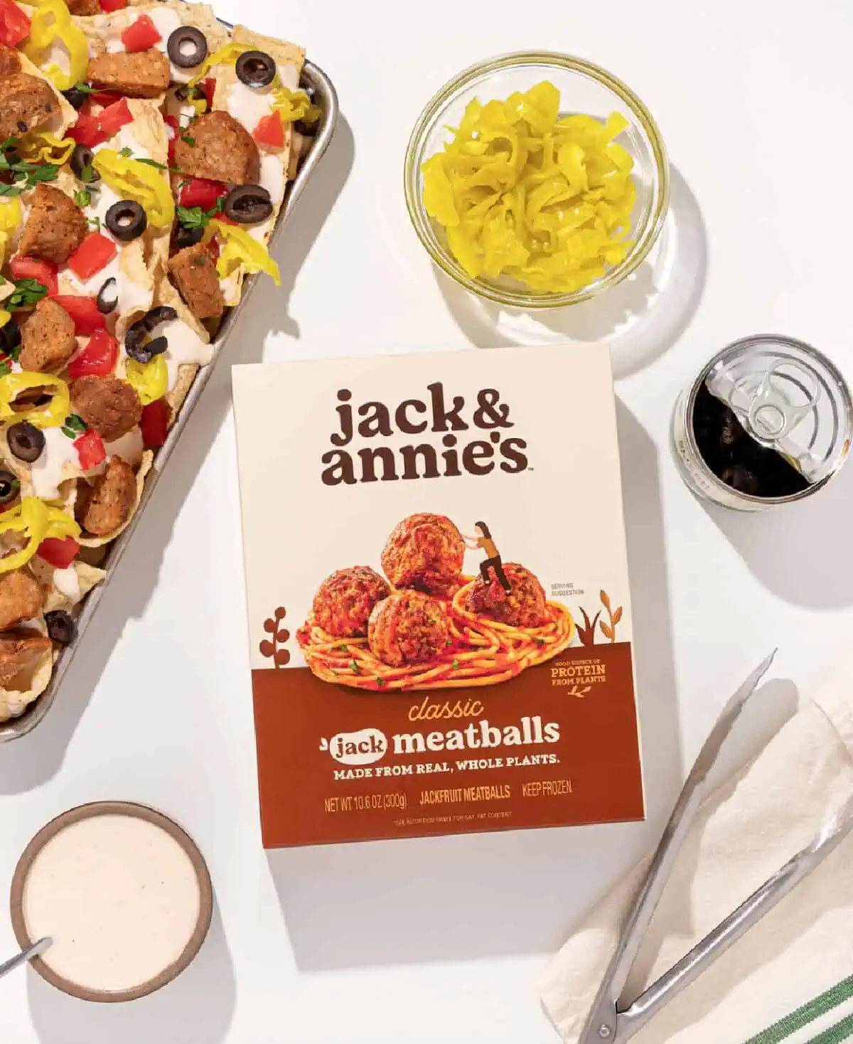 A box of Jack & Annie's classic jackfruit meatballs on a white countertop next to a sheet of nachos, sliced hot peppers, an open can of beans, and a set of silver tongs.