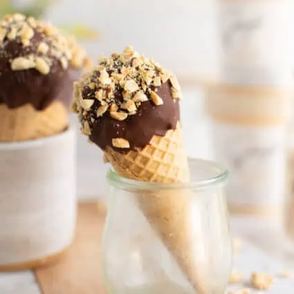 Vegan Drumstick dipped in chocolate and topped with chopped peanuts with pints of ice cream behind it.