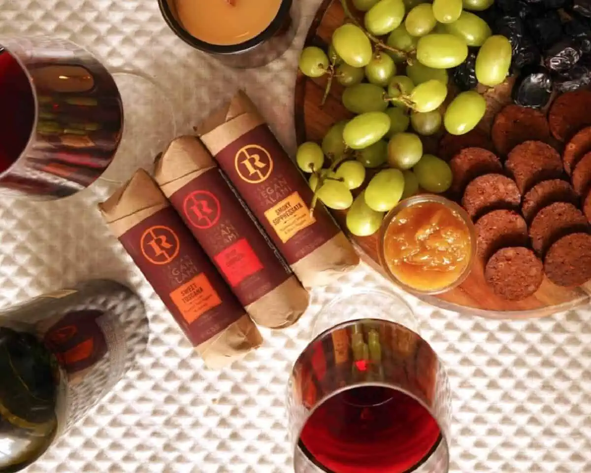 Three cylindrical packages of vegan Renegade Salami surrounded by fruit, glasses of wine and dips on a white patterned table.