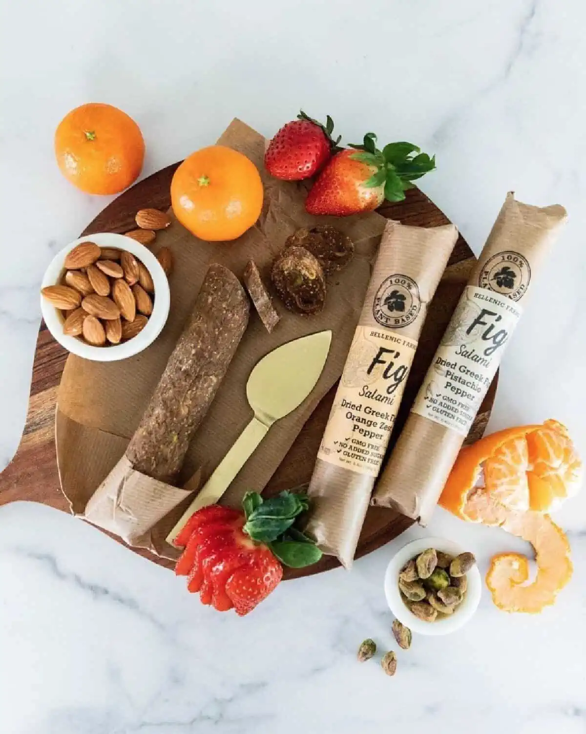 A wooden charcuterie board on the white background displaying various fruits, bowls of nuts, and two flavors of Hellenic Farms vegan fig salami.