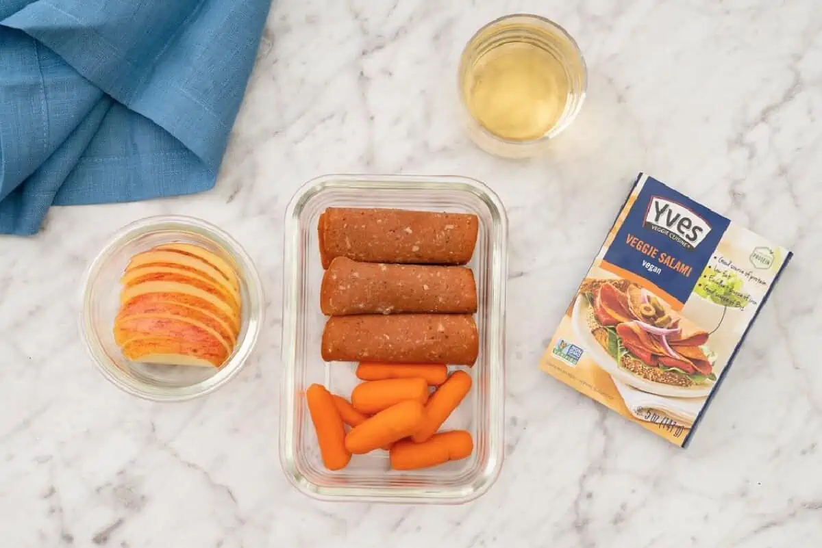 A marble-like tabletop featuring a glass of apple juice, a glass bowl of apple slices, and a glass container holding Yves Veggie Cuisine salami slices alongside baby carrots for a kids lunch.