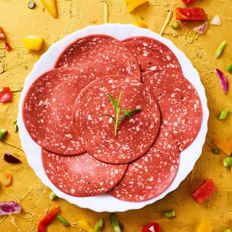 A plate of vegan salami on a yellow background surrounded by diced veggies.