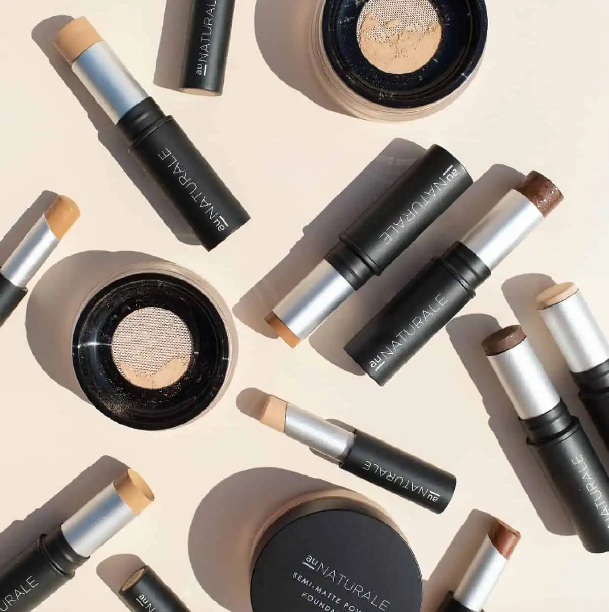 An assortment of Au Naturale vegan makeup products scattered on a light peach background, including their stick foundation and concealer.