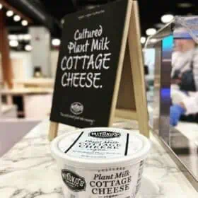 A round white tub of Mikyoko Creamery's vegan cottage cheese on a white marble table top in front of a mini blackboard with the words, "Cultured Plant Milk Cottage Cheese" written on it.