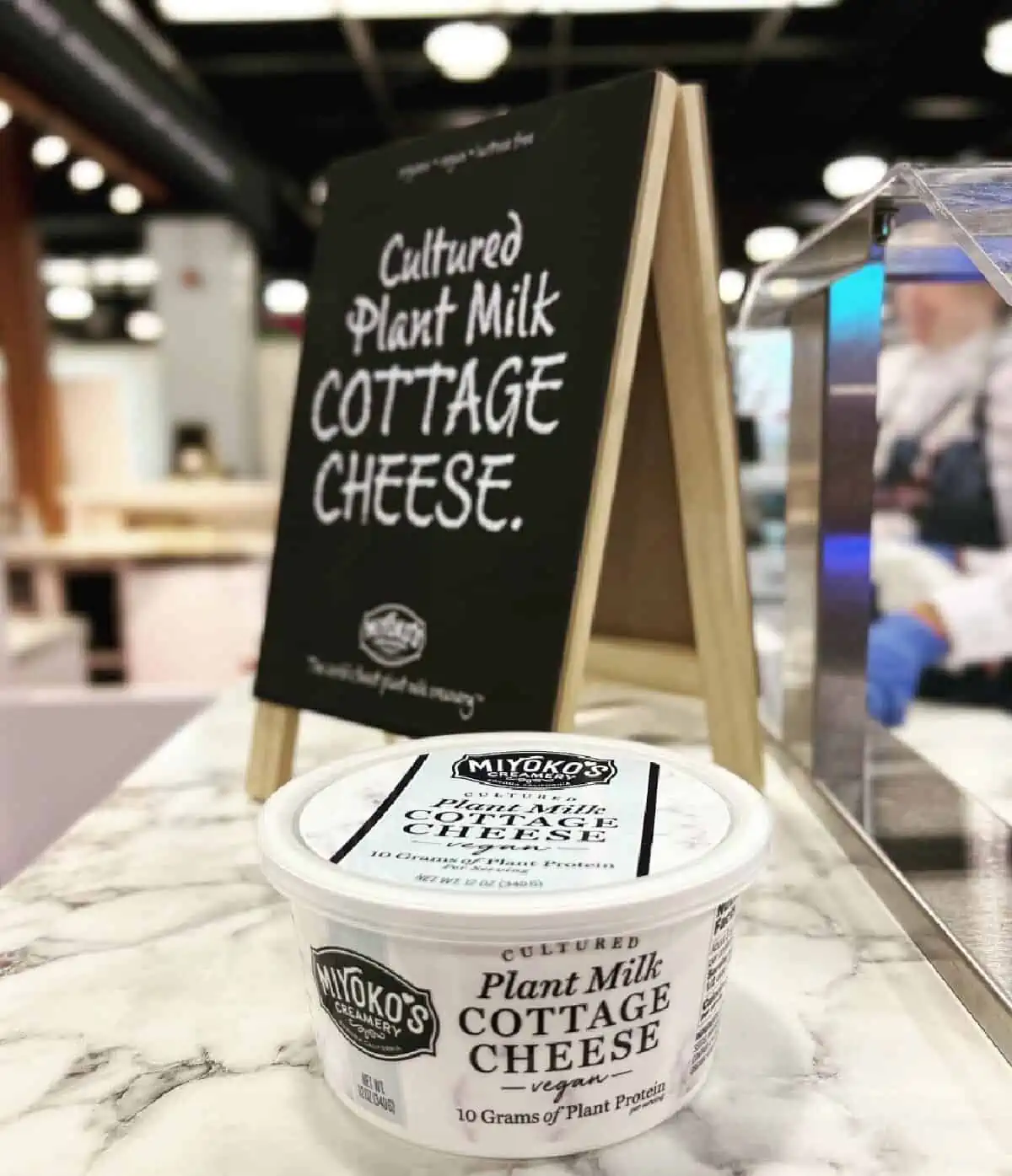 A round white tub of Mikyoko Creamery's vegan cottage cheese on a white marble table top in front of a mini blackboard with the words, "Cultured Plant Milk Cottage Cheese" written on it. 