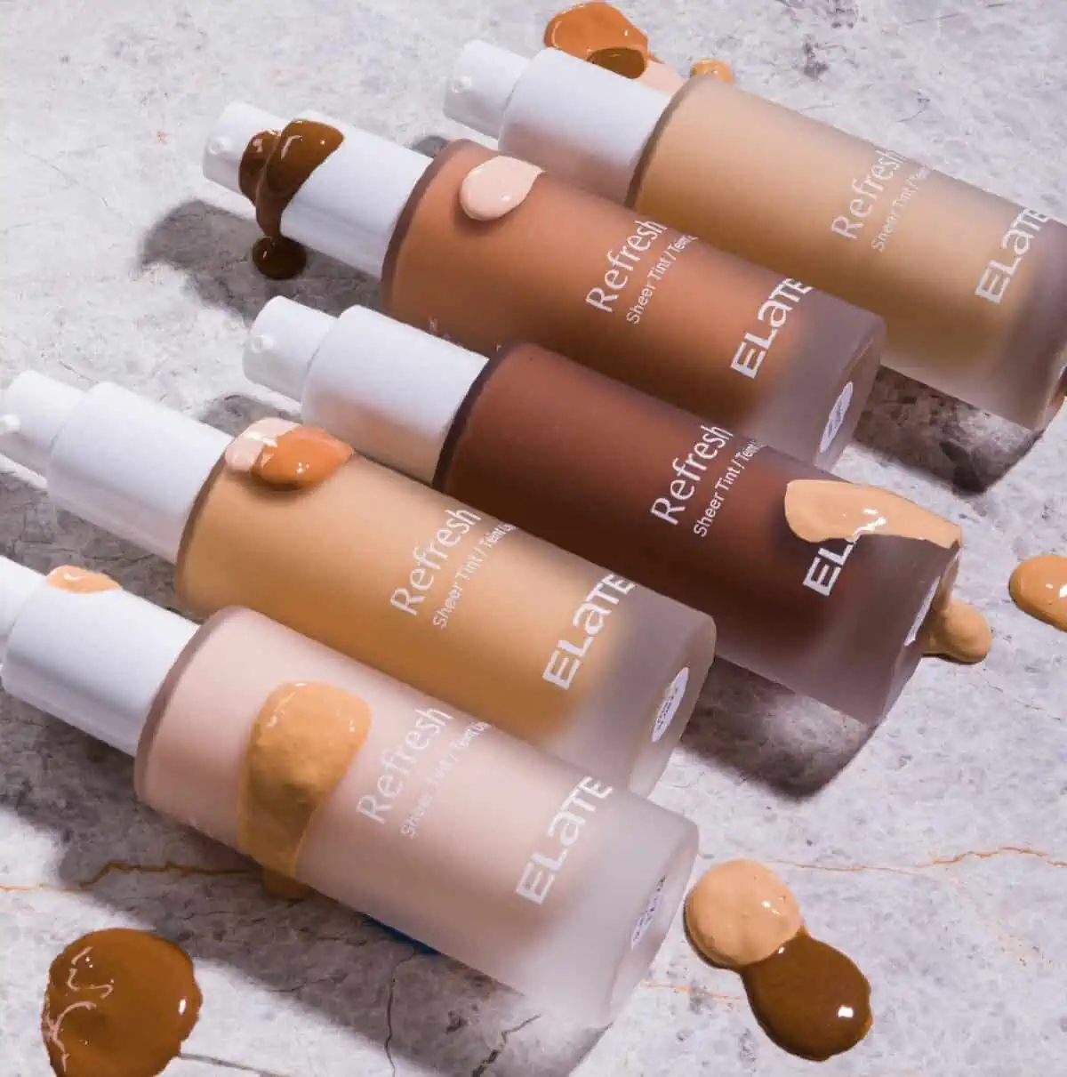 Five bottles of Elate Cosmetics liquid foundation laying on their side on a marble background accented by drops of foundation samples.
