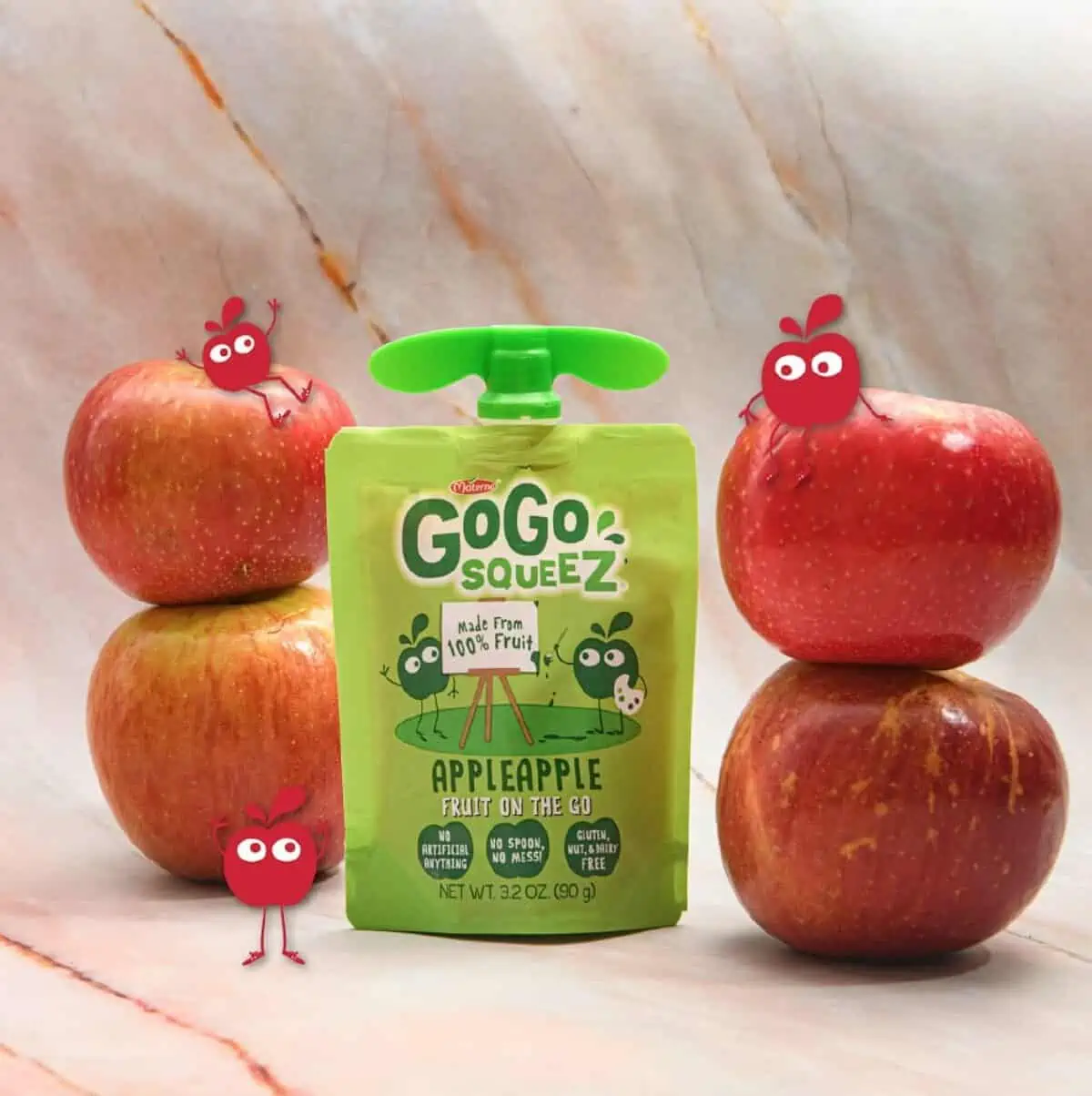 A green Gogo Squeez applesauce pouch standing between two piles of red apples with cartoon characters of apples around them.