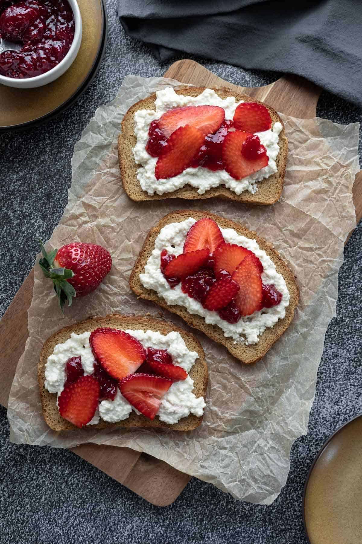 A cutting board covered with wax paper displaying three slices of bread topped with vegan cottage cheese and berries.