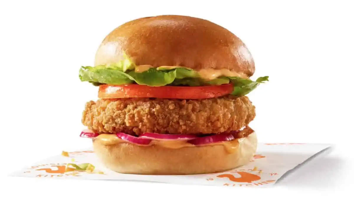 A Popeyes vegan chicken sandwich with lettuce, tomato and creole sauce sitting on a branded square of paper against a white background.