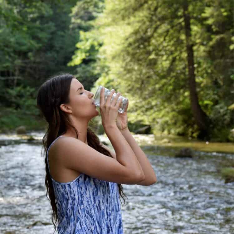 Woman drinking a glass of fresh water in a forest wondering if water is vegan.