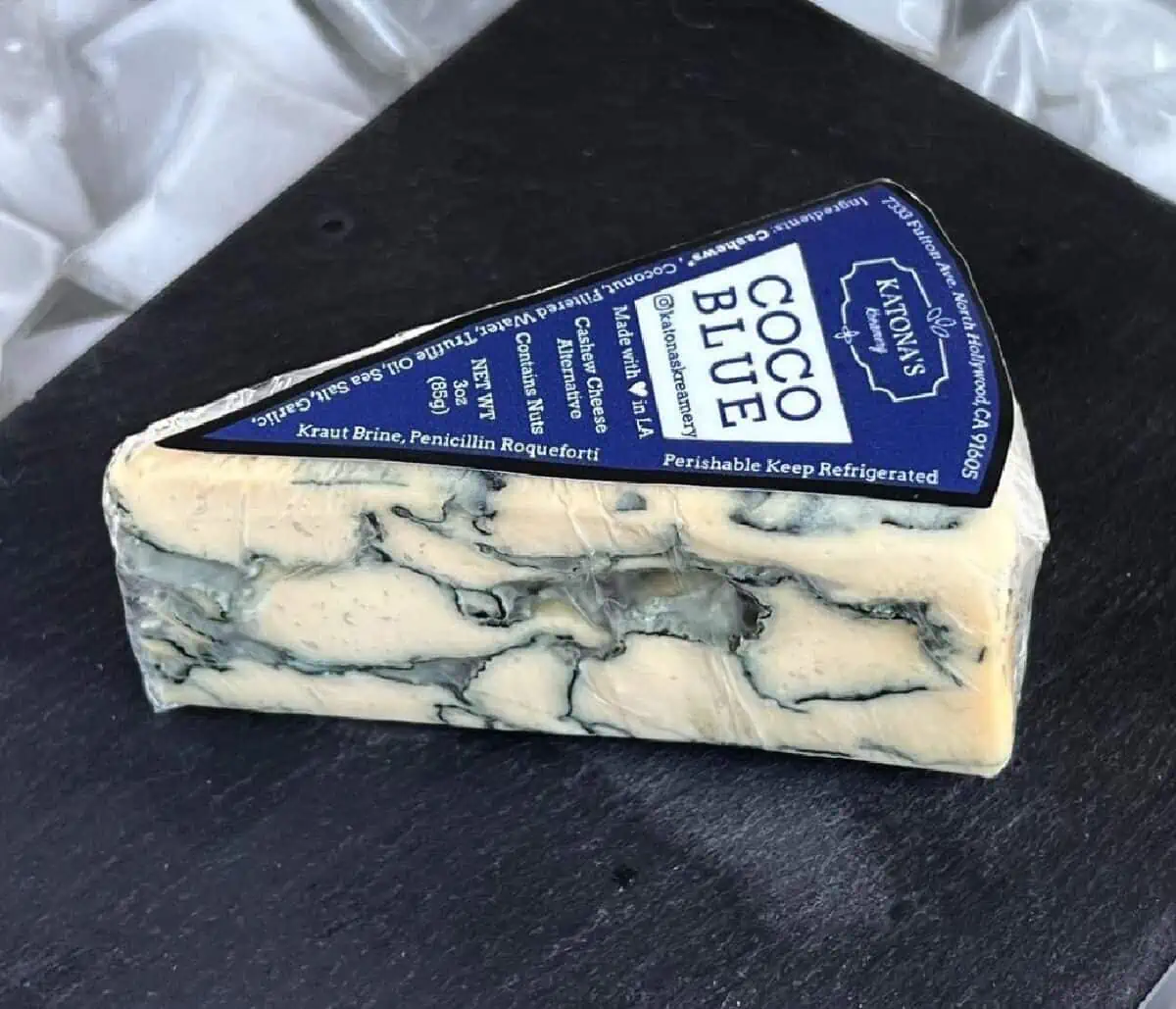 A black plate on top of a white napkin holding a pie shaped wedge of Katonas Kreamery Blue Cheese with dark blue and white label.