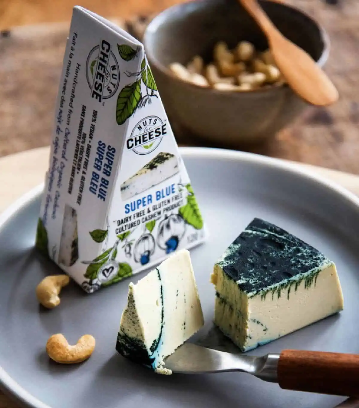 A white plate on a wooden table holding a pie shaped upright wedge of Nuts for Cheese vegan blue cheese along with a slice cut out with a small cheese knife and a few cashews.