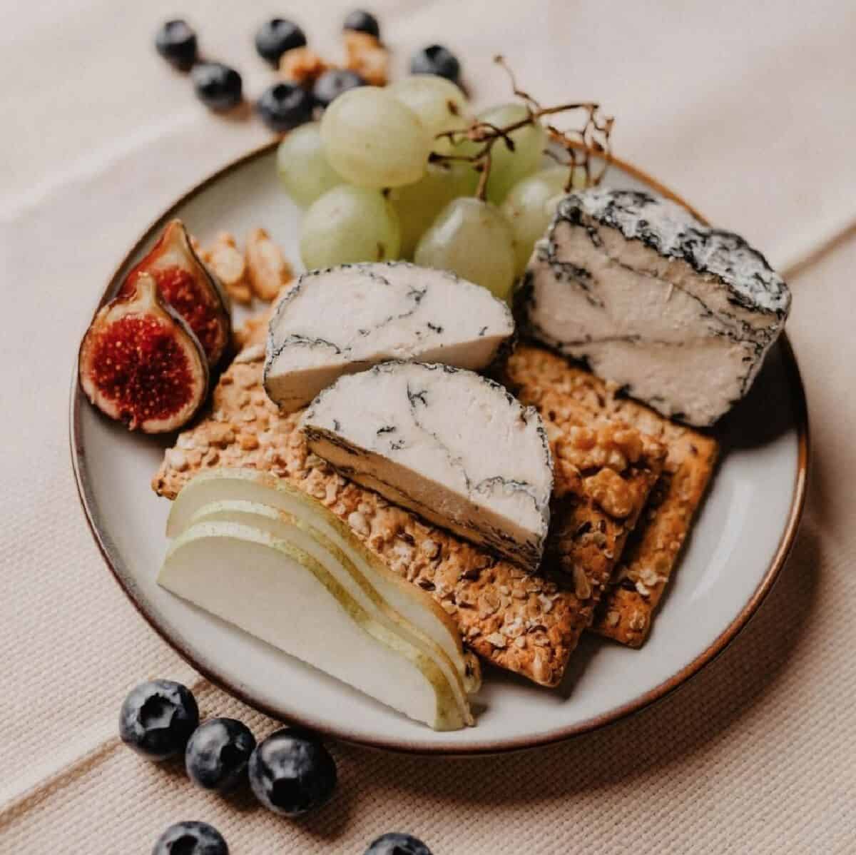 An off white plate holding chunks of Nutty Blue Cheese along with dried and fresh fruit and crackers on a beige table cloth. 