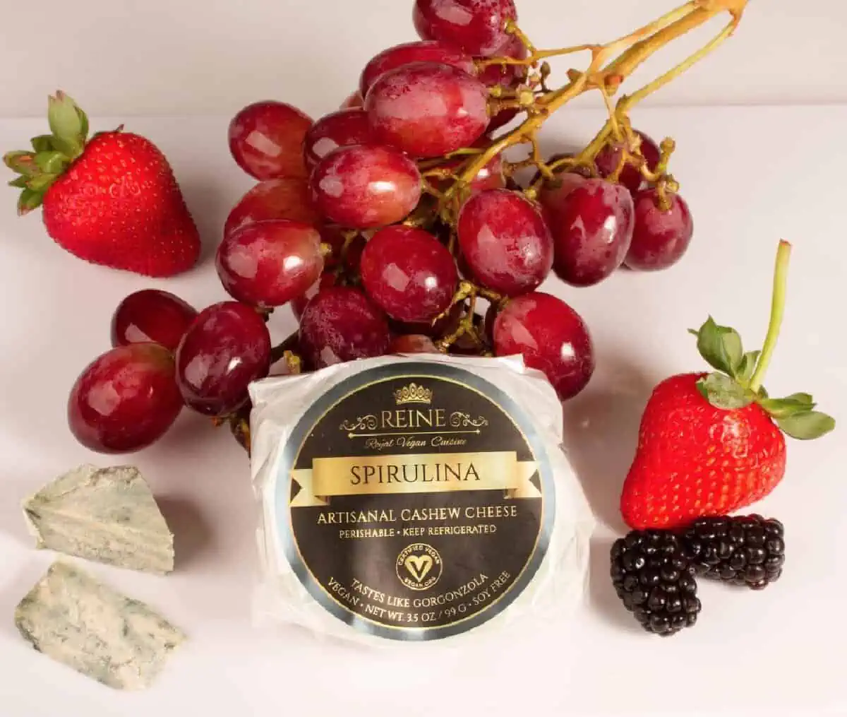 A light colored table holding a white wax paper round of Reine Vegan Spirulina Blue Cheese with a black and gold label surrounded by red grapes, strawberries, and blackberries.