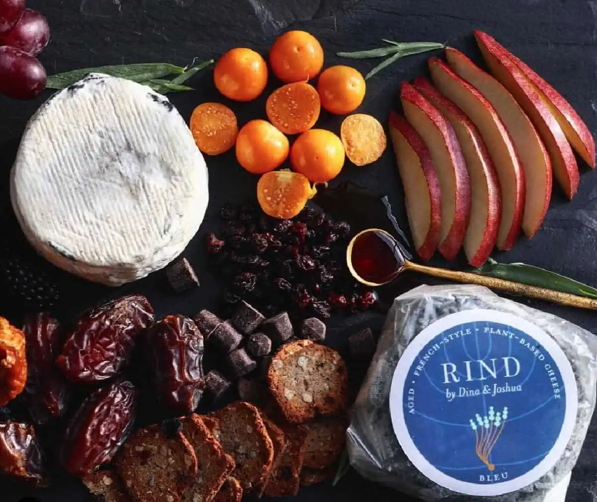 A dark background covered with dried fruits, apple slices, kumquats, and a circle of Rind Bleu Cheese, one covered and one uncovered. 