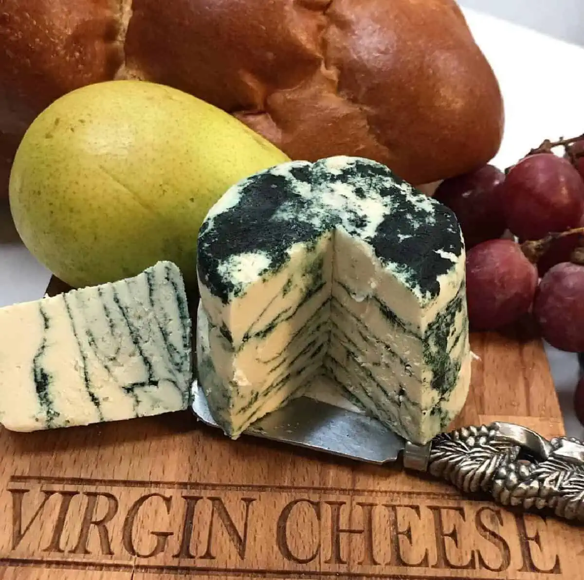 A cutting board with Virgin Cheese etched into the surface holding a cut wedge of vegan blue chese, loaves of bread, grapes, and a pear along with a decorative cheese knife.