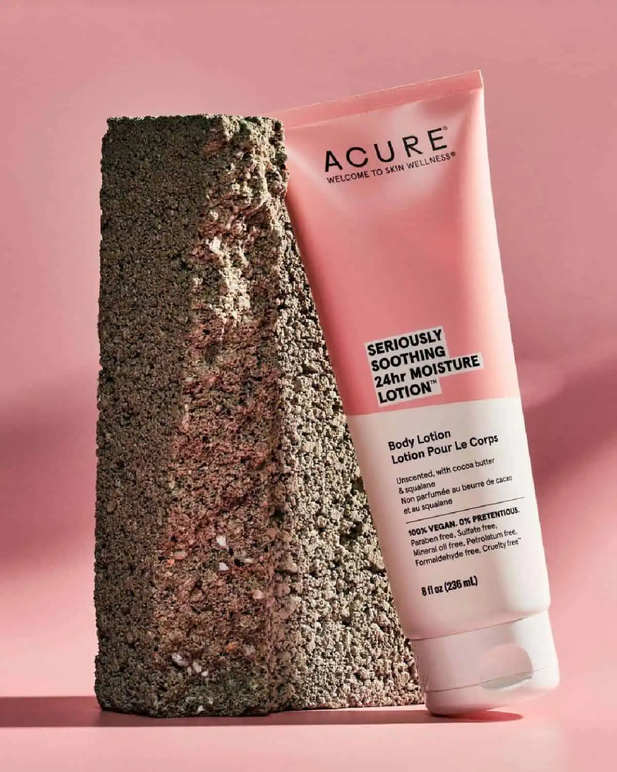 A pink and white tube of Acure body lotion laying against a large rough stone on a pink background.