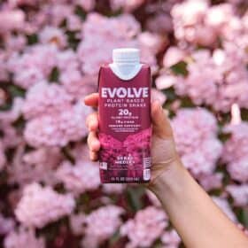 A hand holding a raspberry red colored Evolve plant-based shake container in Berry Medley against a background of pink flowers.