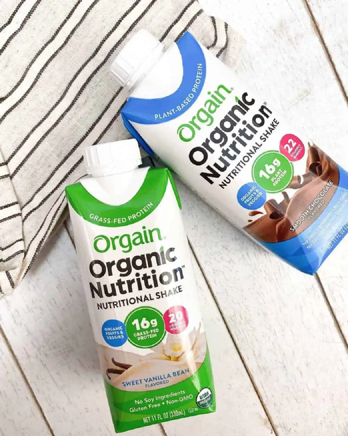 Two Orgain Organic nutrition shakes in vanilla and chocolate flavors leaning against a striped pillow on the light wood background.
