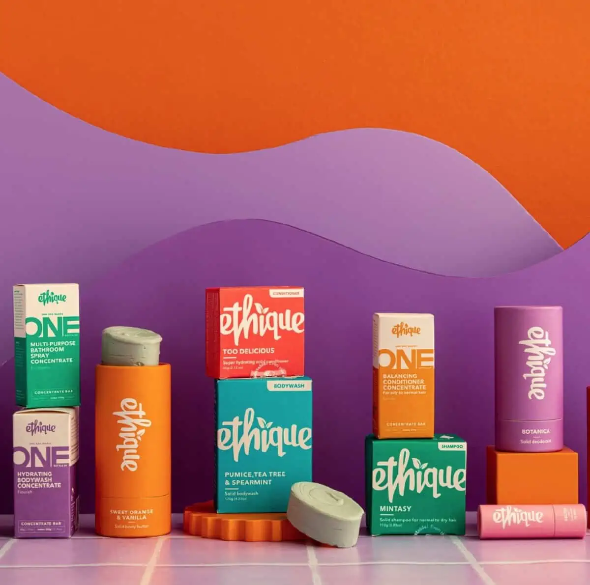 A line up of assorted Ethique vegan and eco-friendly products with a purple and orange patterned background. 