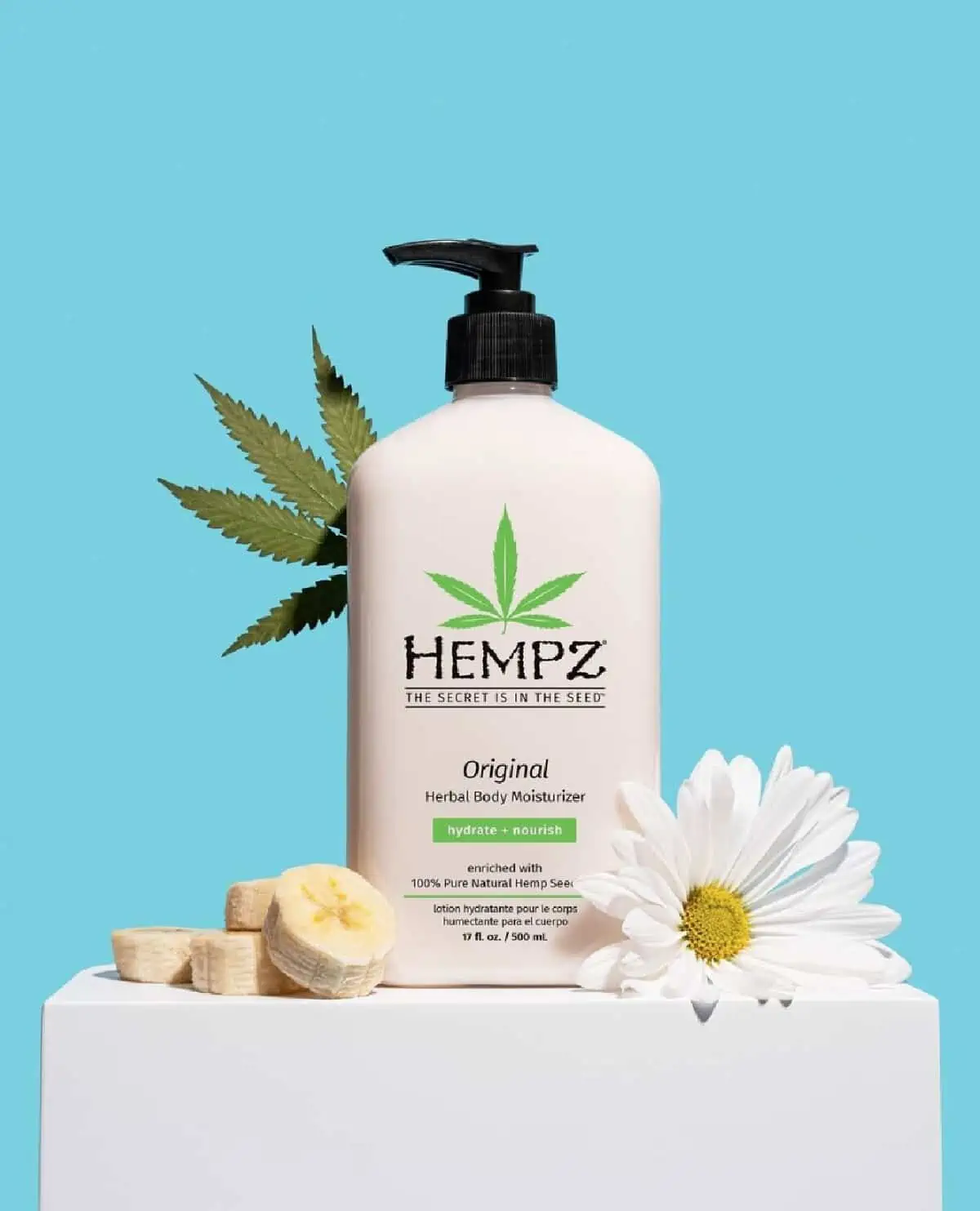 A white bottle of Hempz body lotion with a black pump top on top a white box decorated with bananas, daisies, and a hemp leaf on a blue background. 