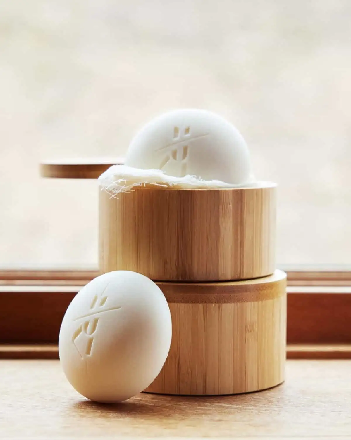 A stack of two round bamboo containers holding two Kate McLeod solid lotion bars against a bright window. 
