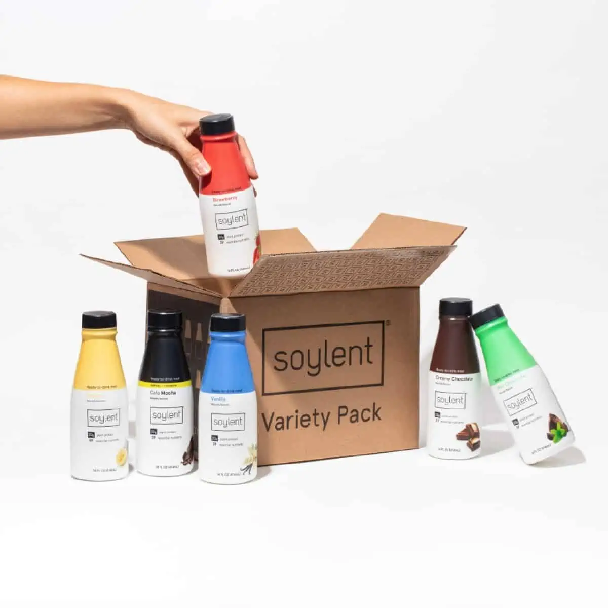 A hand placing a bottle of Soylent plant-based ready-to-drink shake into a cardboard box with five other shake flavors next to the box.