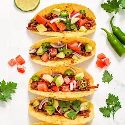 Beefy vegan tacos lined up, served in hard shell tortillas with various ingredients scattered around.