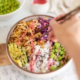 White speckled ceramic bowl with shredded tofu, rice, edamame, avocado, purple cabbage, and pink sauce, and a women's hand scooping up the tofu with chopsticks.