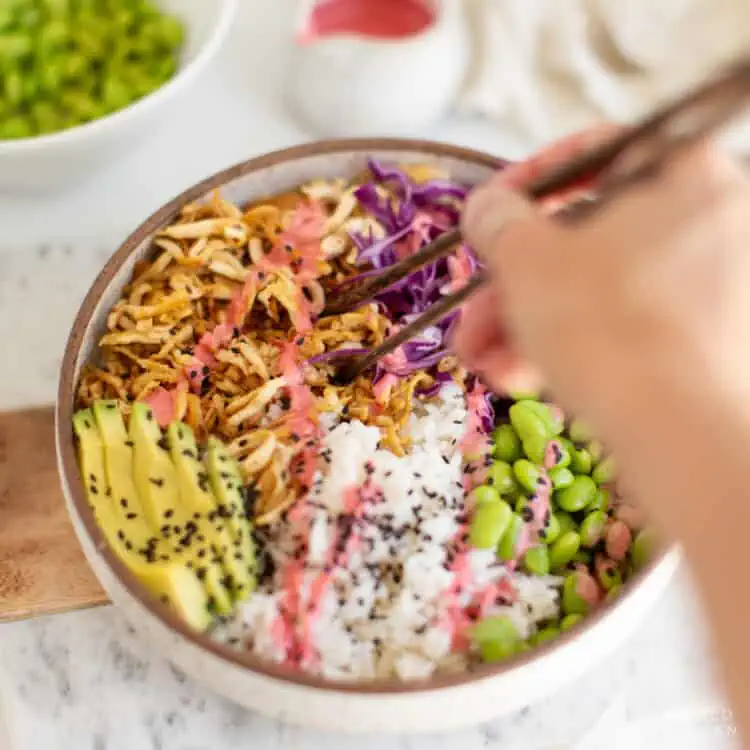 White speckled ceramic bowl with shredded tofu, rice, edamame, avocado, purple cabbage, and pink sauce, and a women's hand scooping up the tofu with chopsticks.