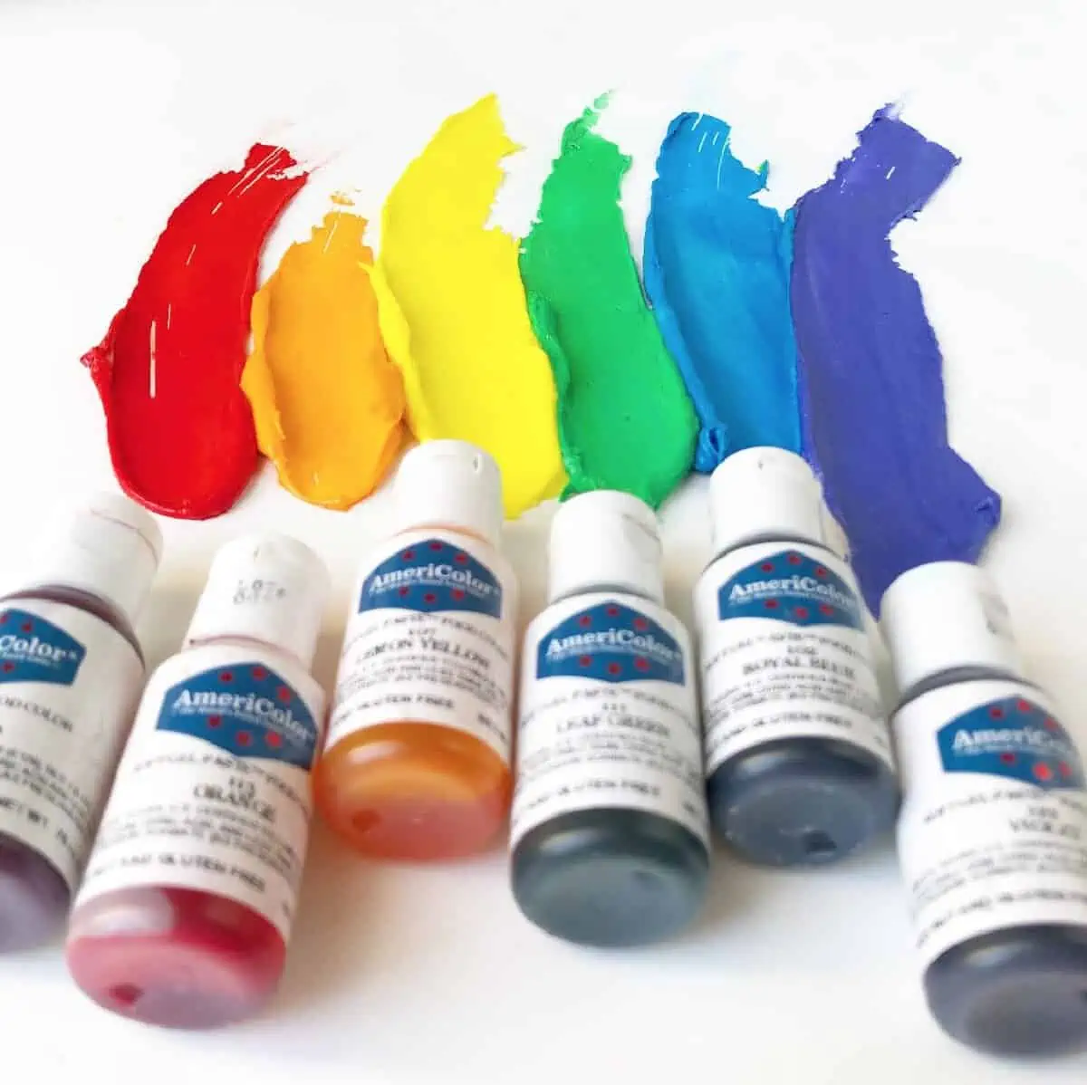 Six plastic bottles of Americolor food coloring paste laying on a white background with samples of rainbow colors streaking above.