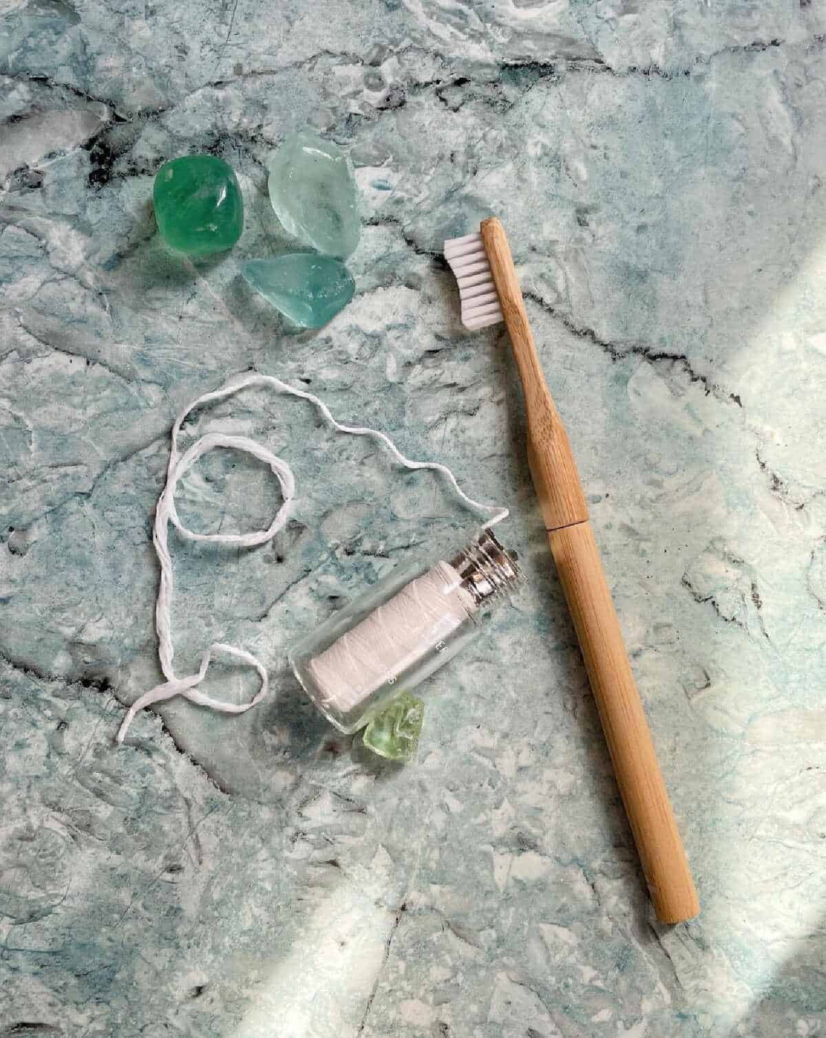 A small glass container full of vegan-friendly floss on a light green marble background and next to a bamboo toothbrush.