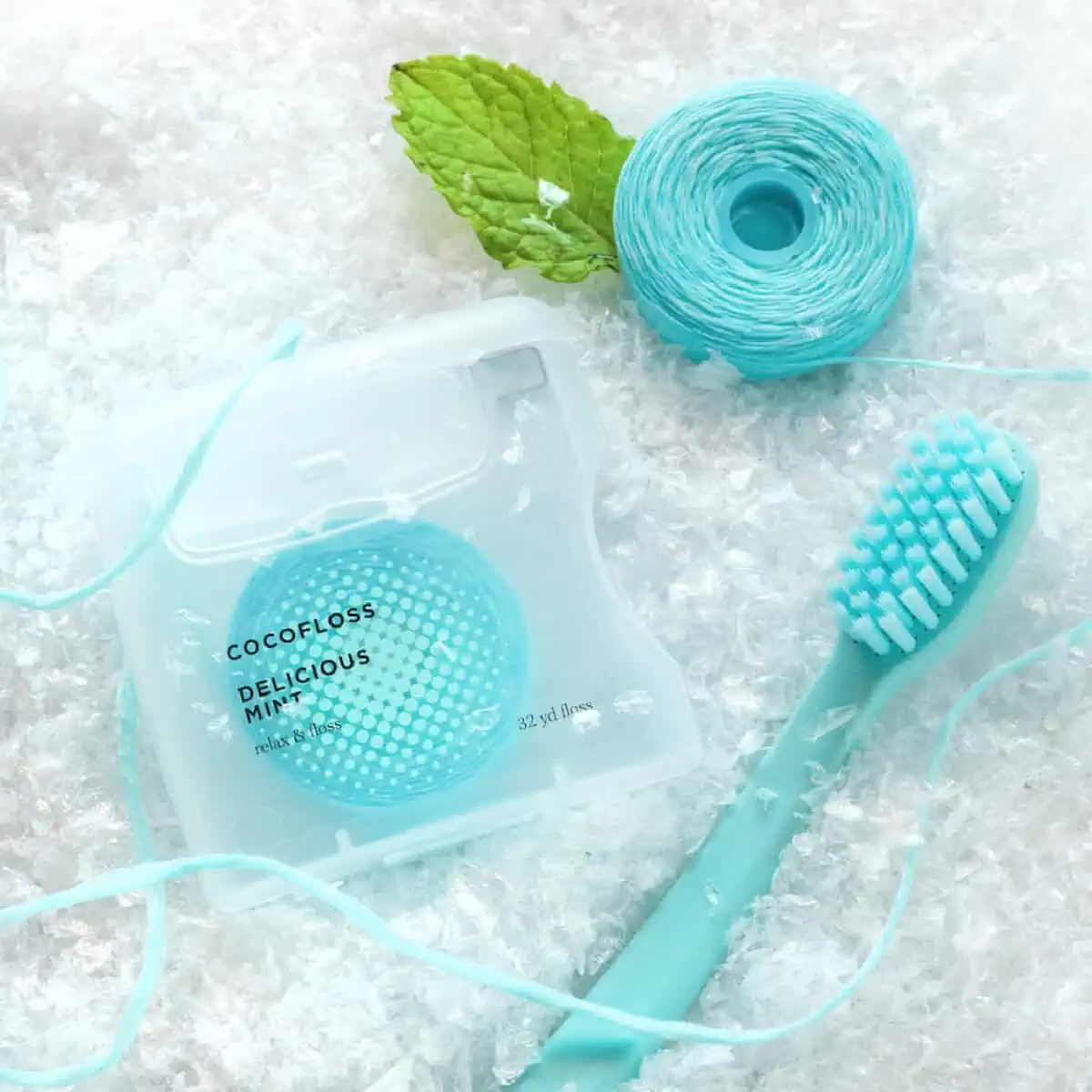 A clear container of turquoise colored vegan Coco Floss on a snowy background next to more floss, a matching toothbrush, and a peppermint leaf.