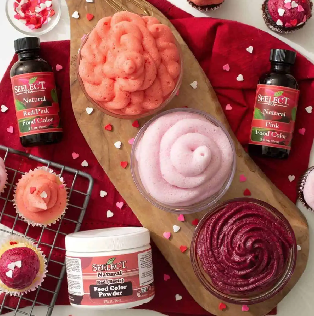 Two bottles and one jar of Nature's Flavors food coloring among cupcakes and other red and pink baked goods on a wooden tray and red napkin.