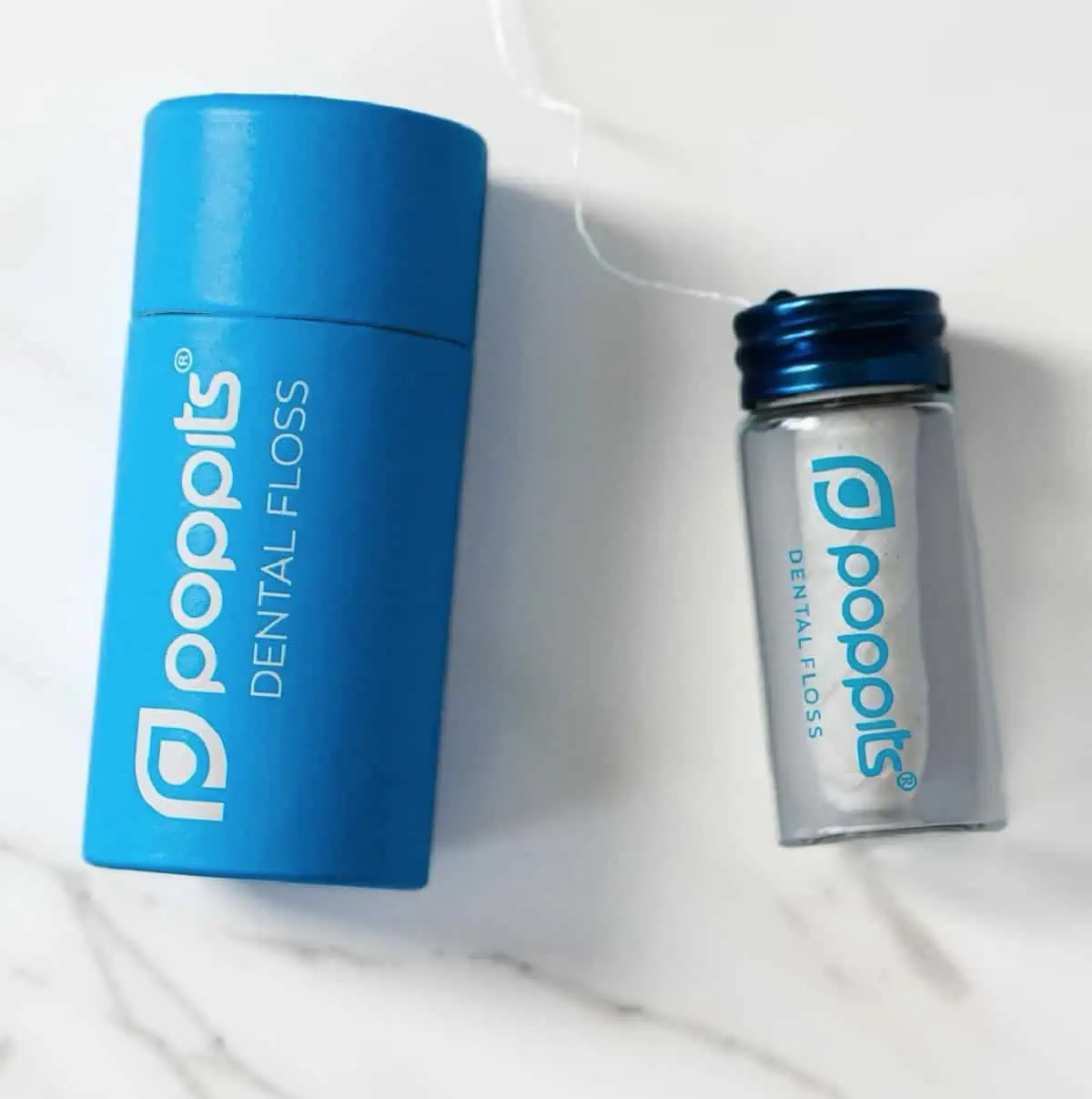 Blue cardboard container and glass floss container for Vegan Poppits Floss on a white marble background.