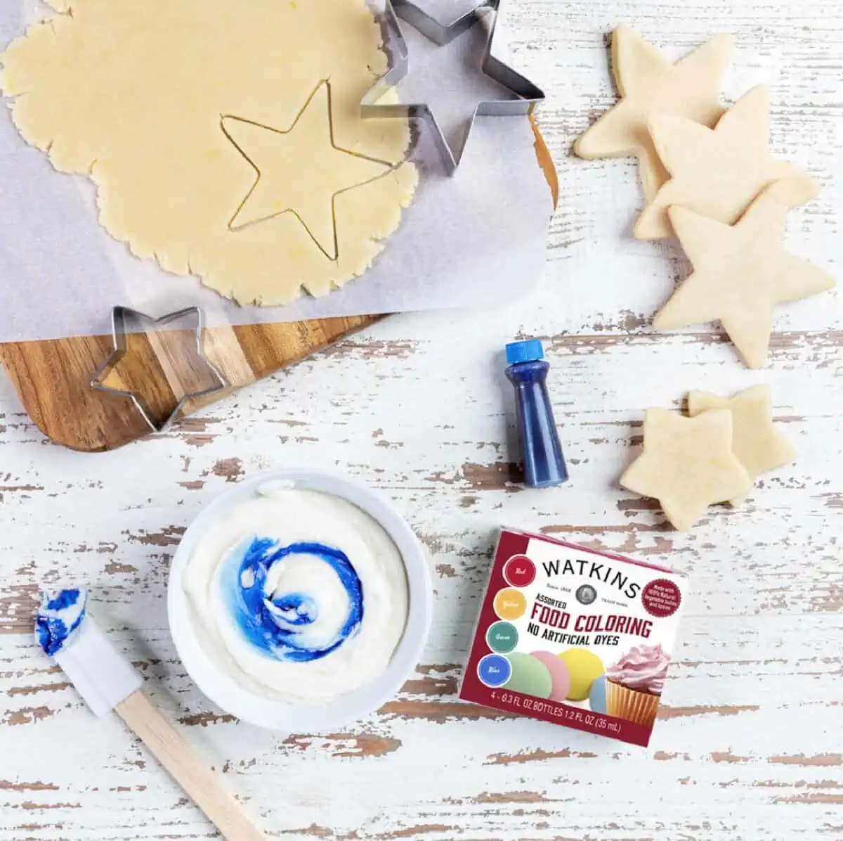 A box of Watkins food coloring on a white wooden background surrounded by uncooked, cut cookies, cookie cutters, and frosting.