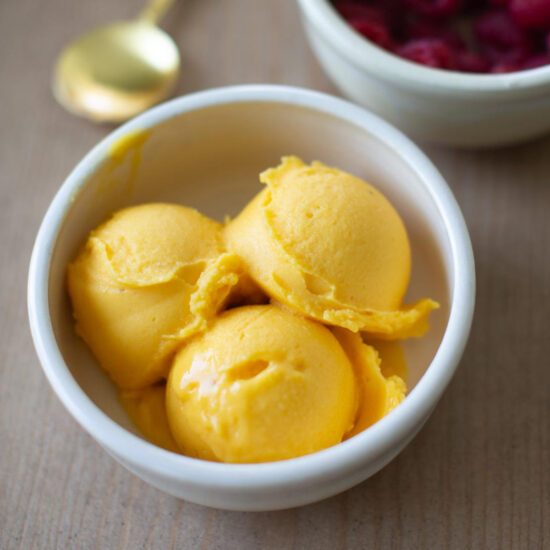 A bowl of mango sorbet with three scoops next to a bowl of fresh raspberries.