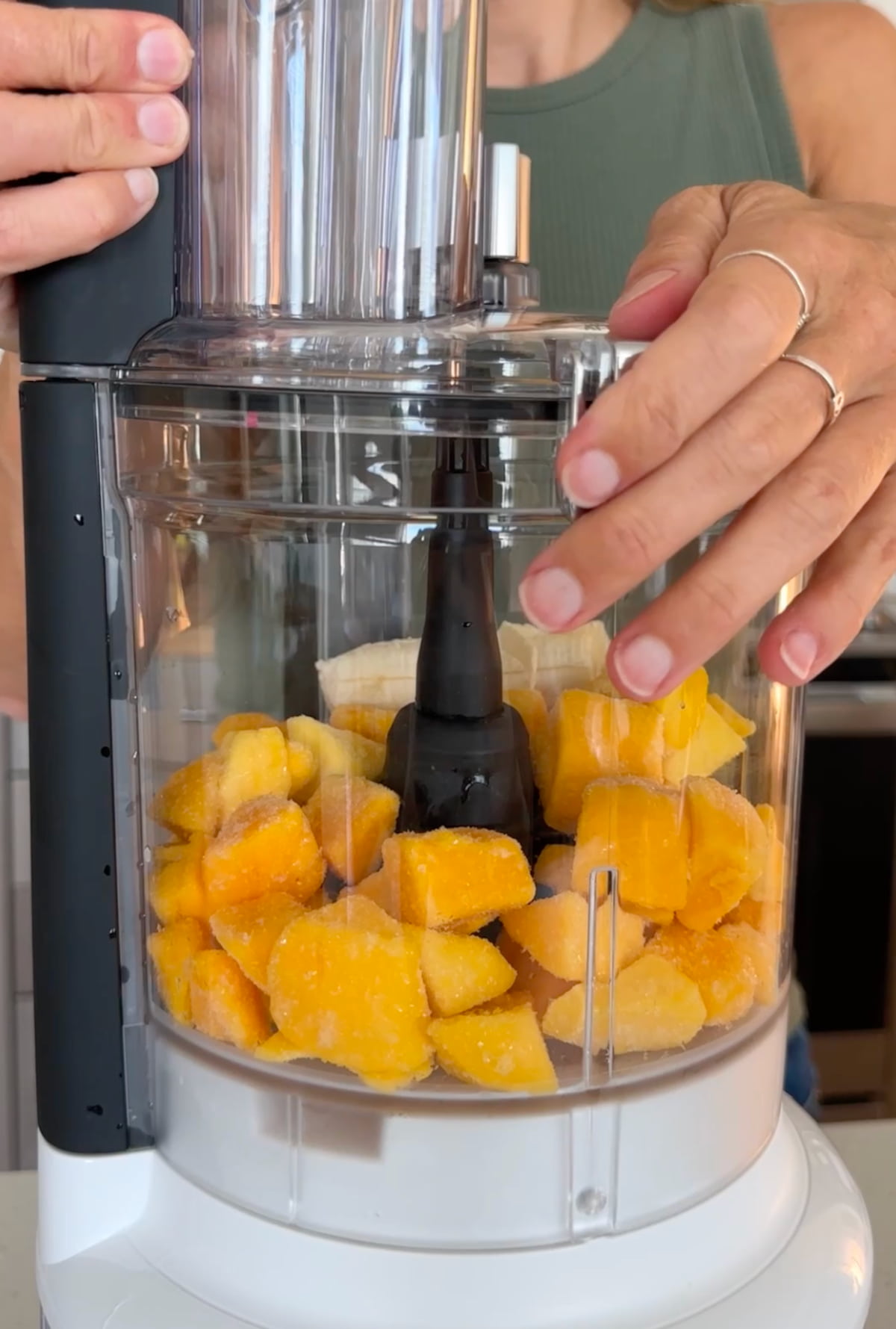 A woman is using a food processor to blend frozen mango and ripe banana together.