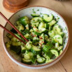 A bowl of spicy asian cucumber salad topped with red pepper flakes and served with chopsticks.