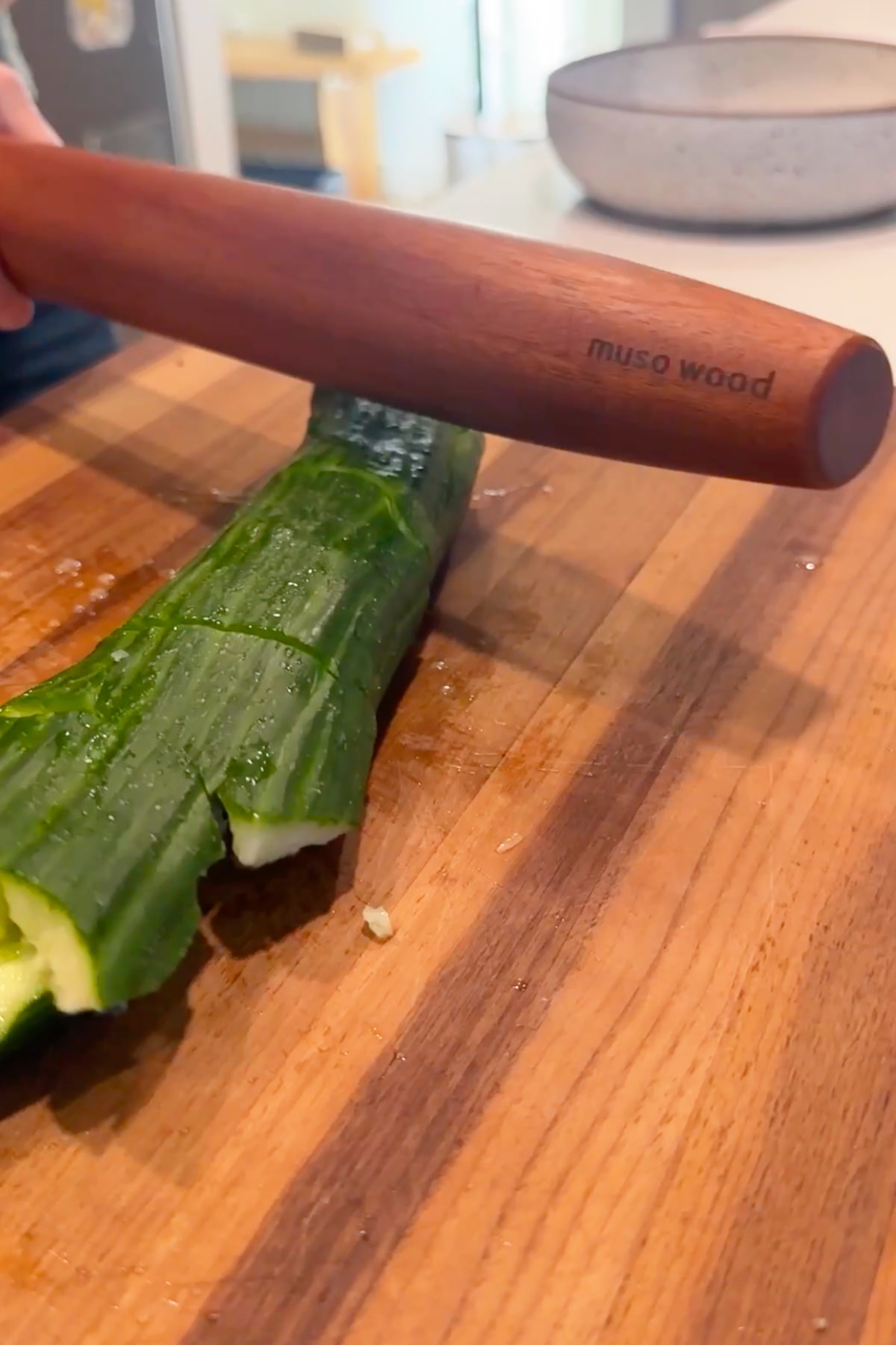 A large cucumber on a cutting board getting smashed by a wooden rolling pin.