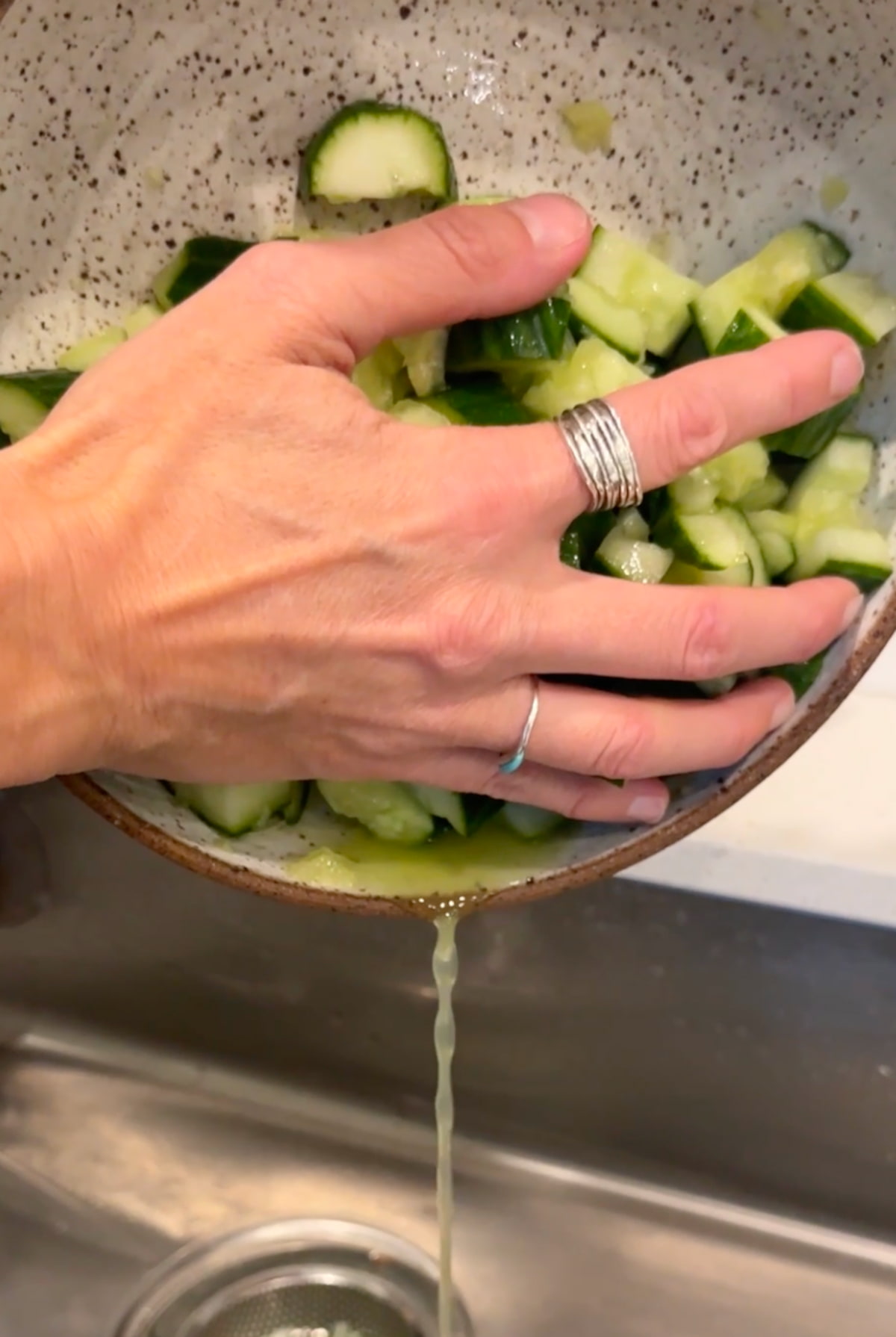 Hands holding a bowl of salted cucumber over a sink to drain the excess liquid.