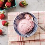 A bowl of vegan strawberry ice cream with fresh strawberries on a marble countertop.