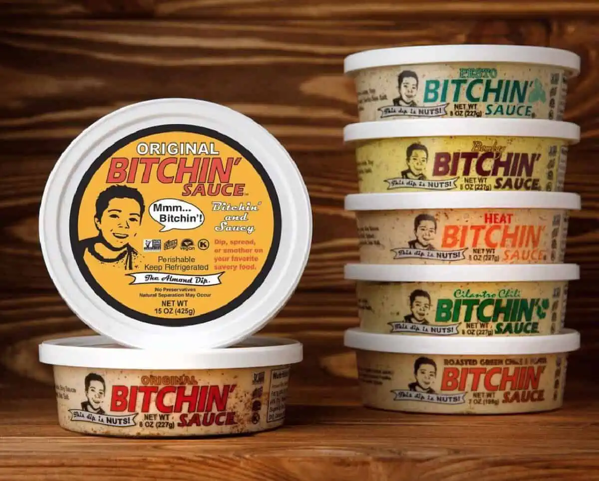 A two stacks of Bitchin' Sauce round containers in various flavors against a wood paneled background. 
