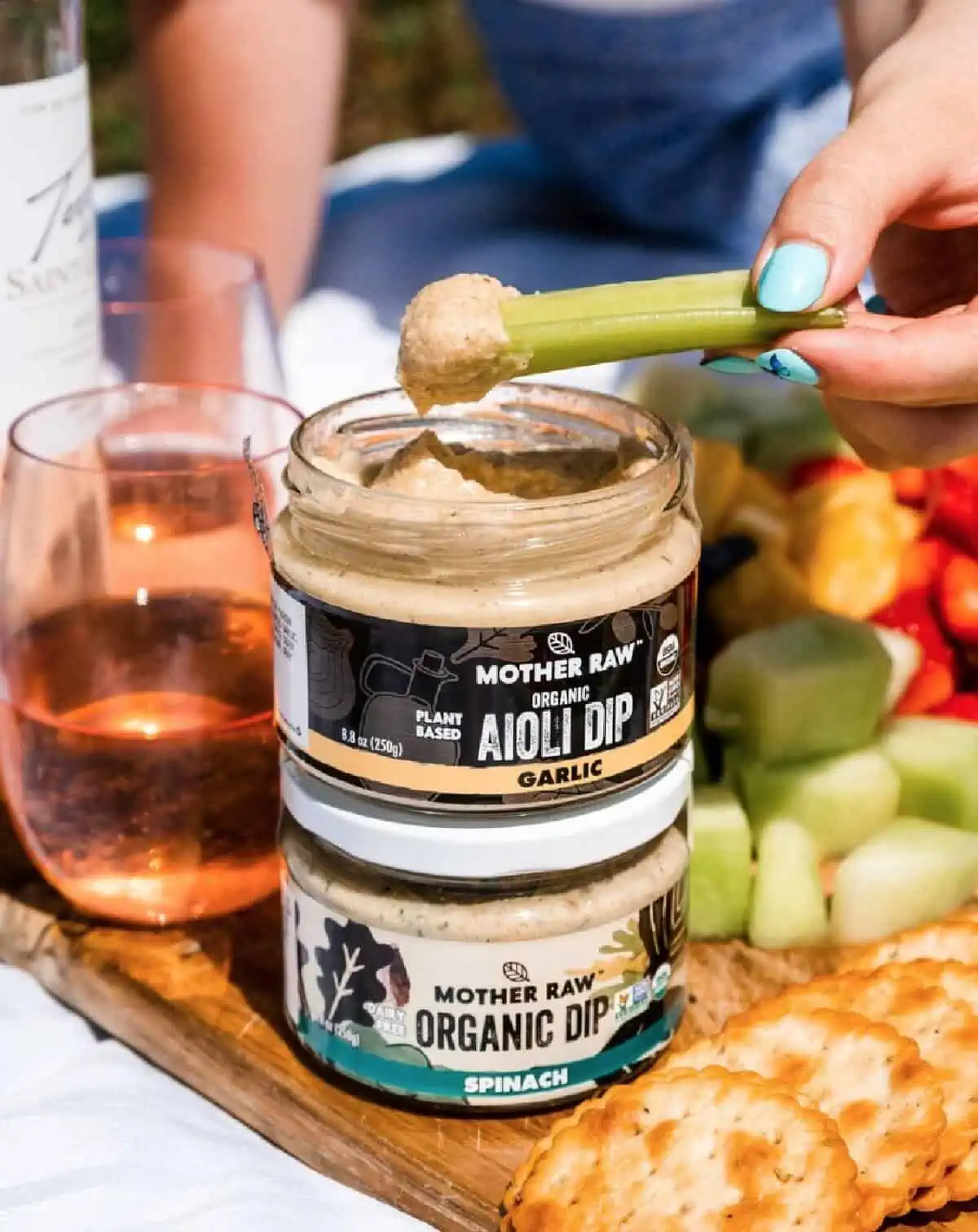 A wooden cutting board filled with vegetables, crackers, and wine along with two jars of Mother Raw plant-based organic dip.