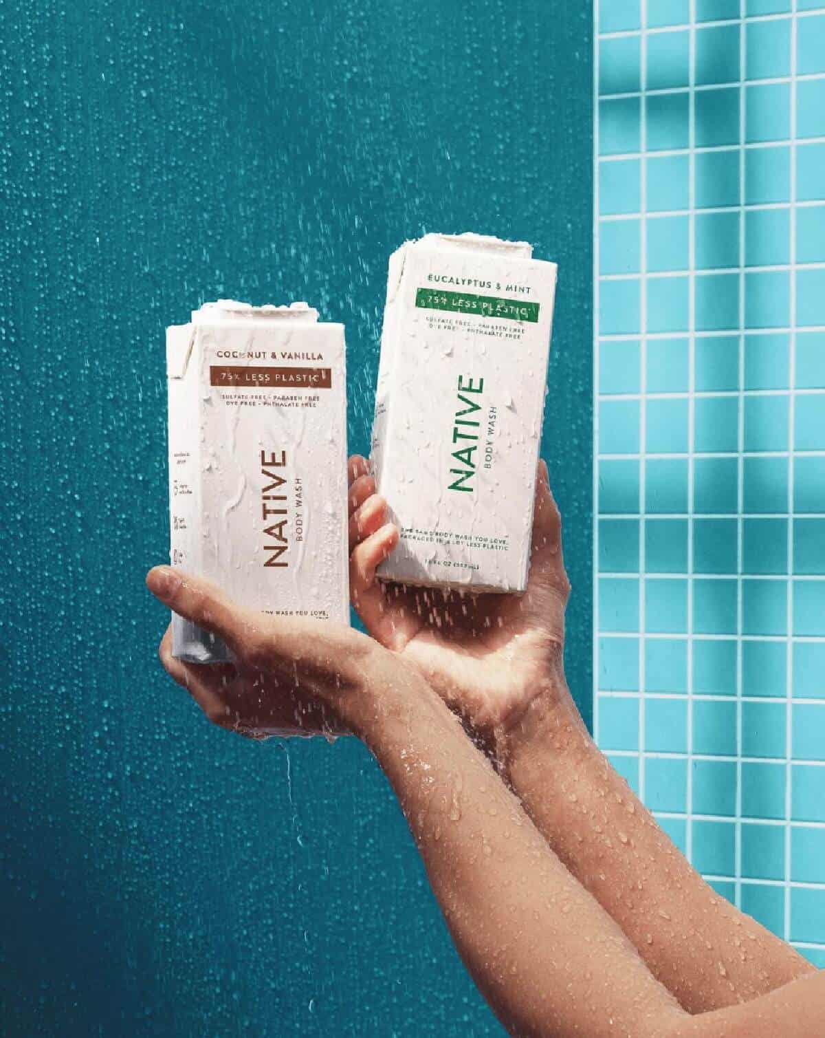 Two hands each holding up a white container of Native body wash against a teal blue shower background. 