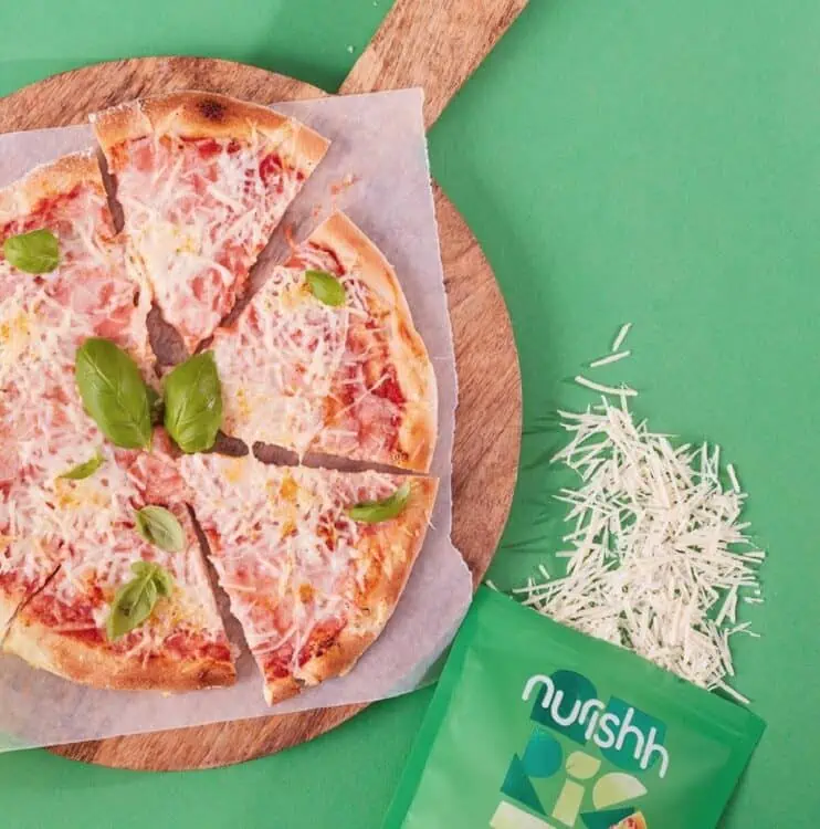 A vegan cheese and basil pizza on a wooden pizza board on a green background, next to a green package of Nurissh plant-based mozzarella cheese.