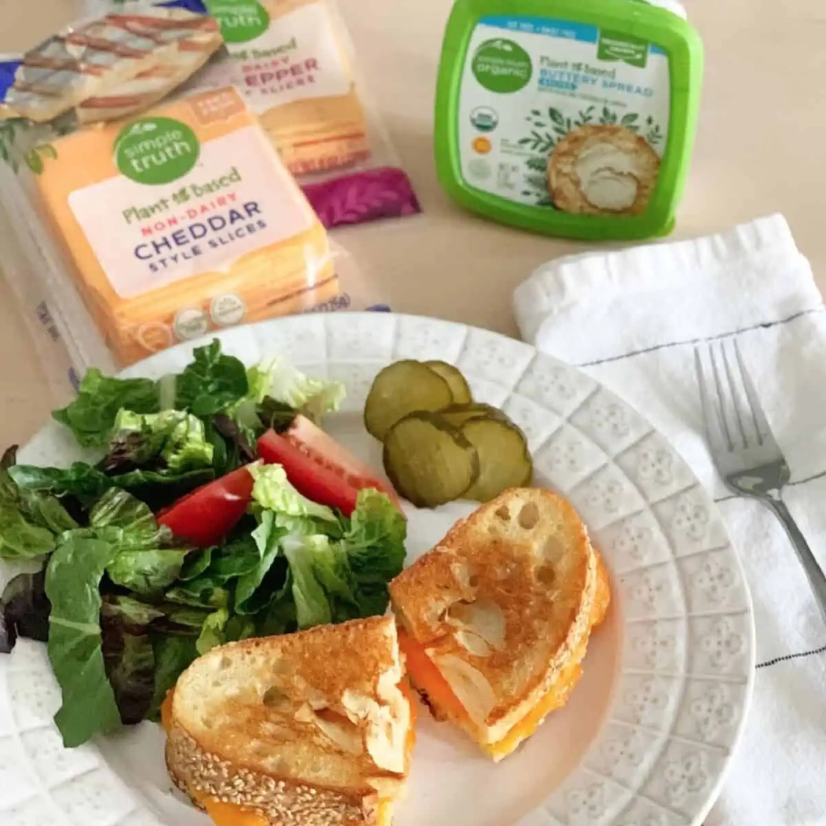 A plate filled with a vegan grilled cheese, salad, and pickles next to a package of Simple Truth dairy-free, plant-based cheese slices and buttery spread. 