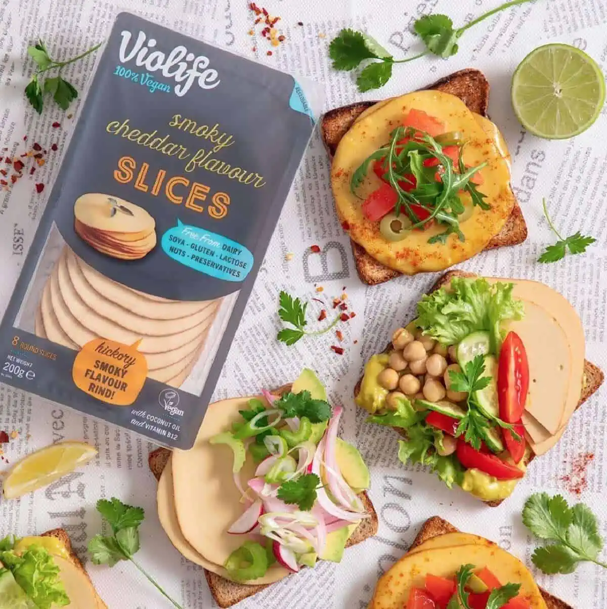 Violife 100% Vegan Cheese Slices next to three plant-based, open-faced toast sandwiches on a newspaper background. 