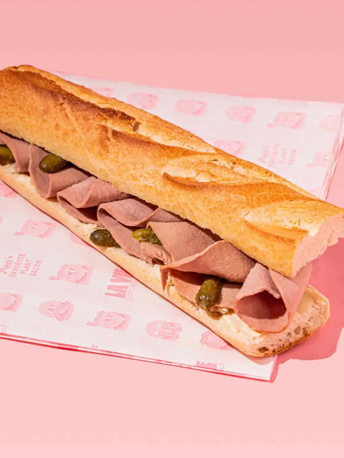 A sandwich with La Vie vegan ham slices folded inside of it on deli paper with a pink background.