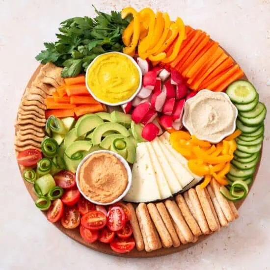 A round vegan charcuterie board filled with chopped vegetables, fruit, crackers, and a few different hummus flavors.